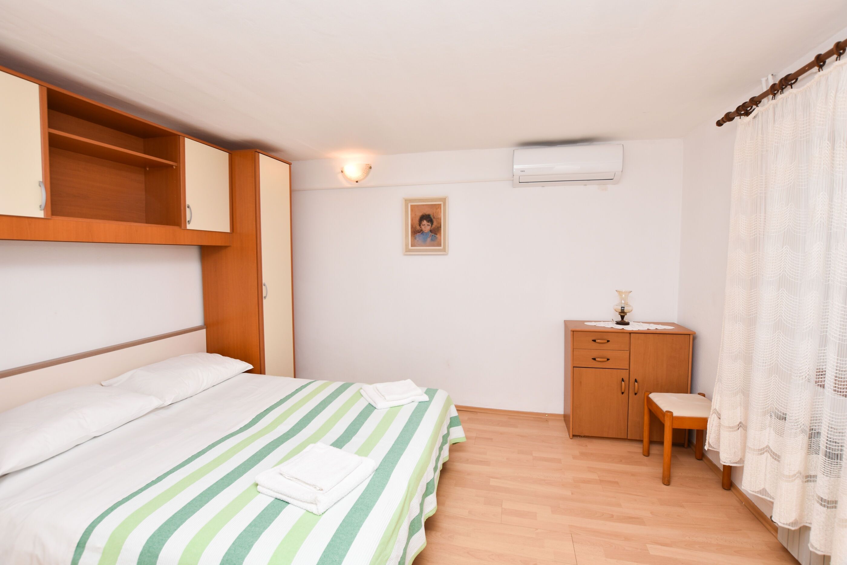 A bedroom with a bed, a wooden wardrobe, a desk with a chair, and a picture on the wall.