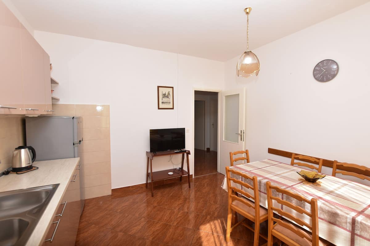 A compact kitchen with a sink, stove, and a television on a stand. There is a dining table with chairs and a clock on the wall.