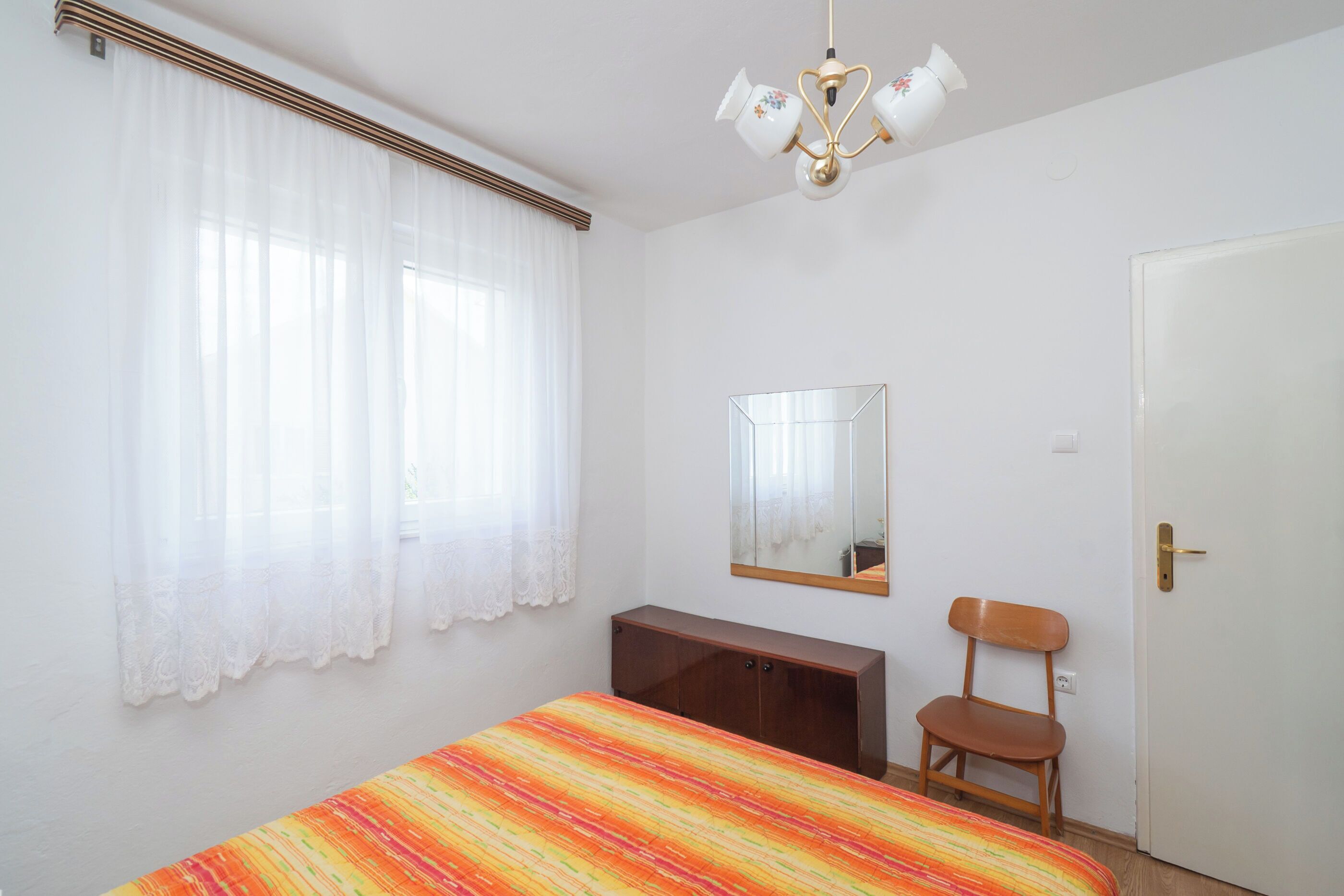 A bedroom with a bed, a wooden chair, a desk, a mirror, and a door.