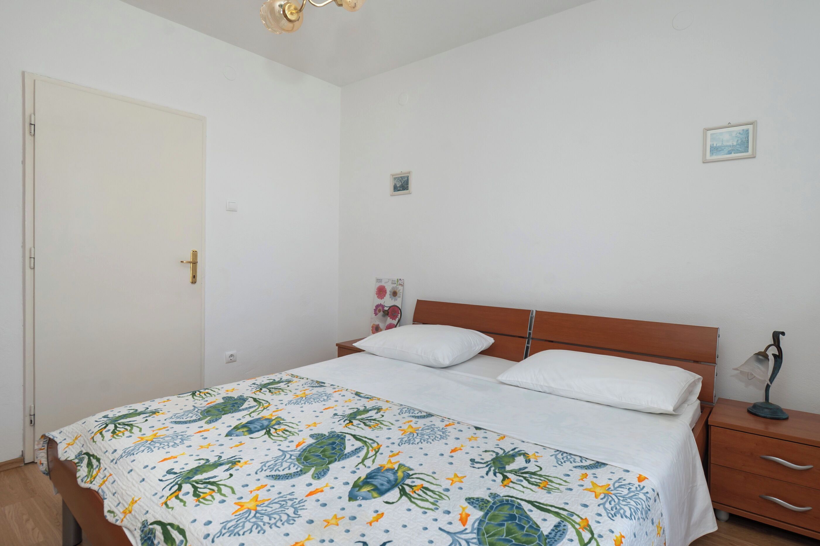 Un dormitorio con cama, mesita de noche, lámpara, una puerta y un cuadro pequeño en la pared.