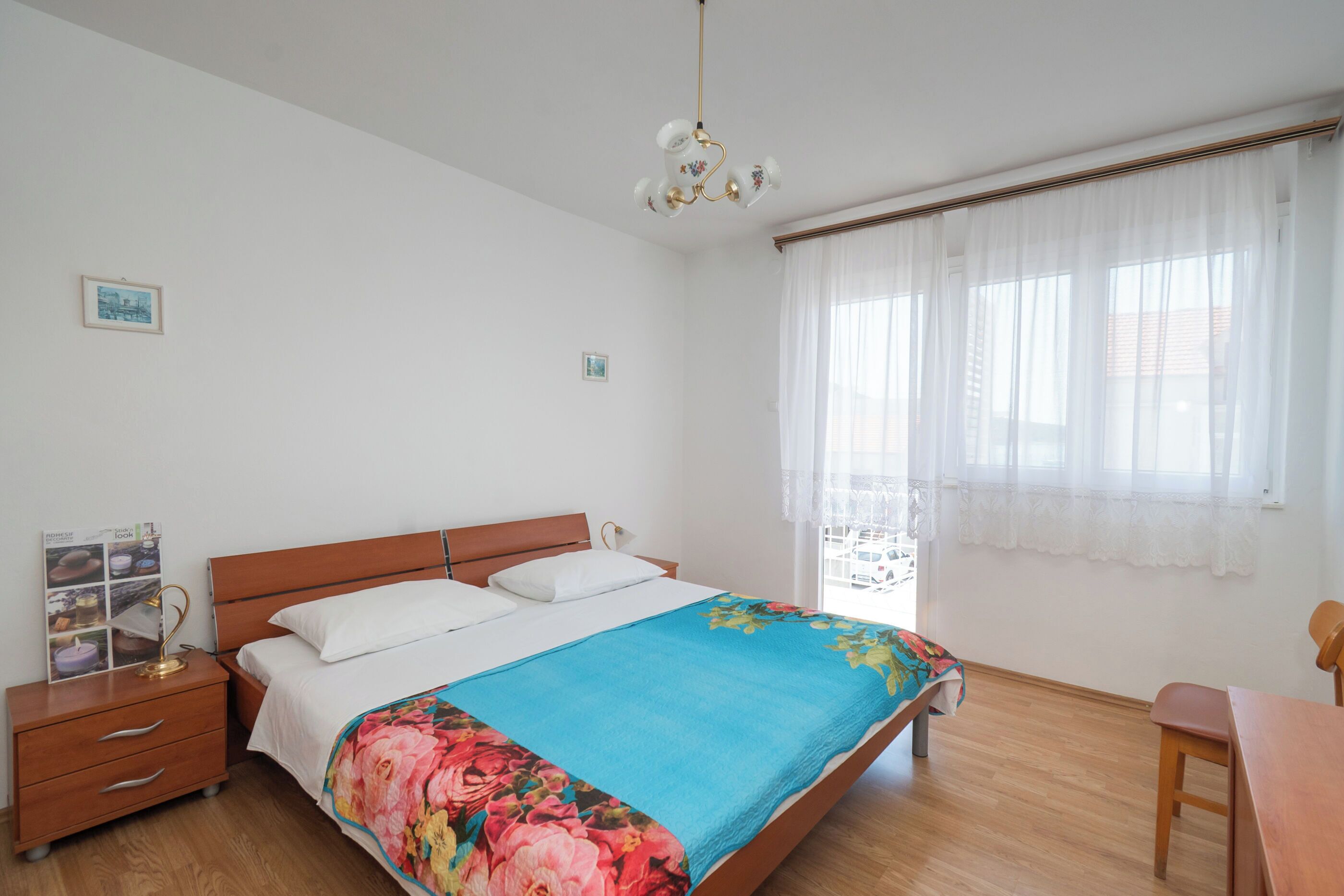 A bedroom with a bed, wooden nightstand, lamp, and a window with sheer curtains.