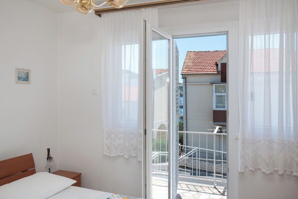 Une chambre à coucher avec un lit, une table de chevet, une fenêtre avec des rideaux blancs et un balcon doté d’une balustrade.