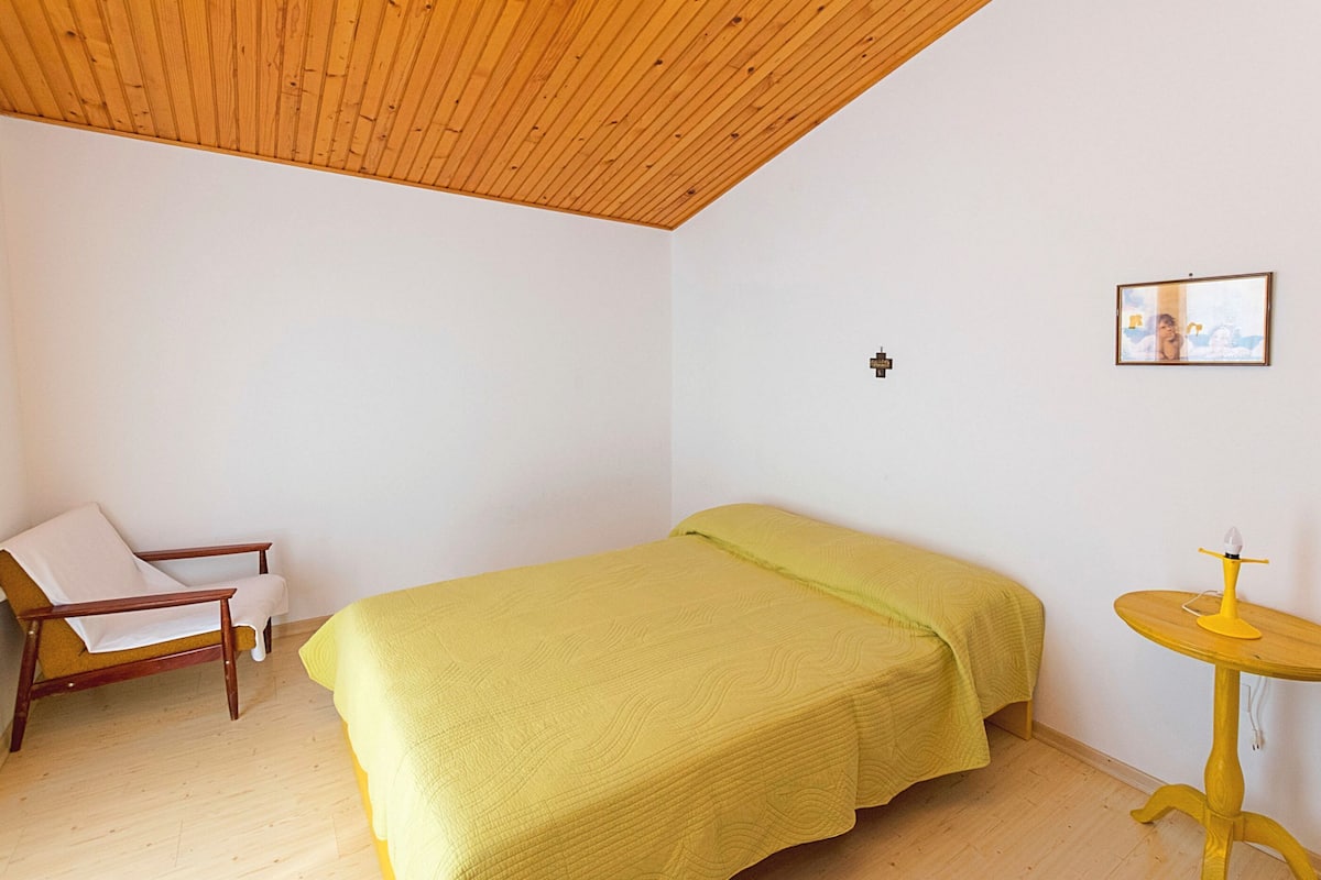 A room with a bed, a chair, a small table, and a wooden ceiling.