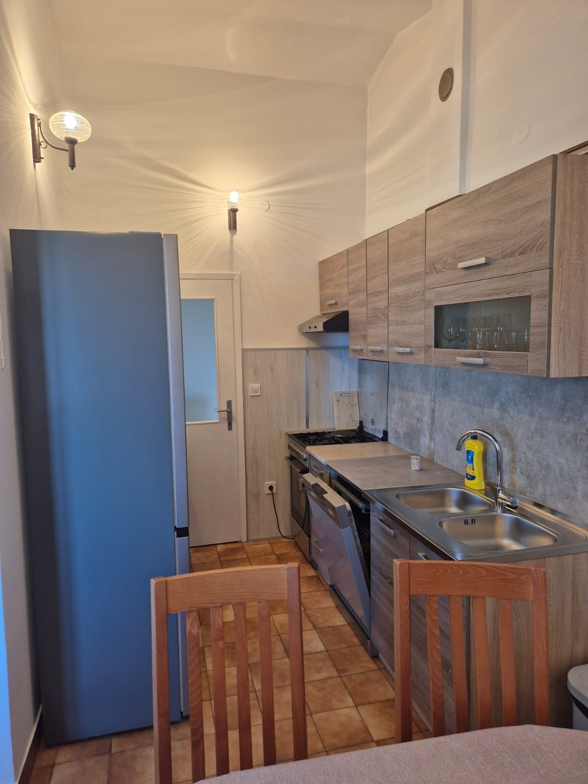 Una cucina compatta con un frigorifero blu, mobili in legno, un lavandino in acciaio inossidabile e un piccolo angolo ristoro con due sedie.