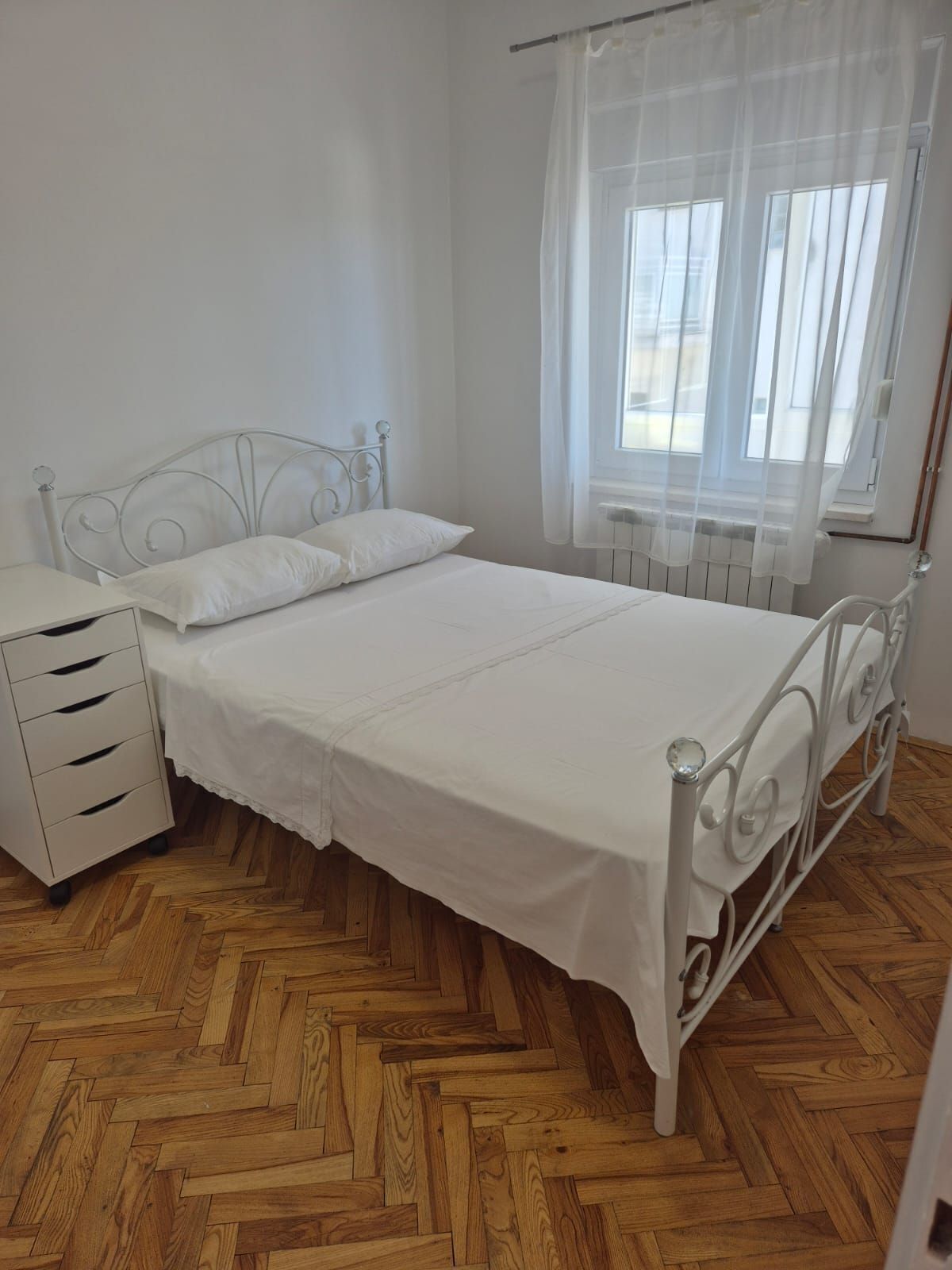 A single-bed room with a metal frame, white bedding, a wooden herringbone floor, a bedside table with drawers, and a window with sheer curtains.
