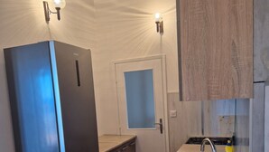 A compact kitchen with a black refrigerator, wooden cabinets, a sink, and two wall-mounted lights. - Apartment Bore (Primosten)