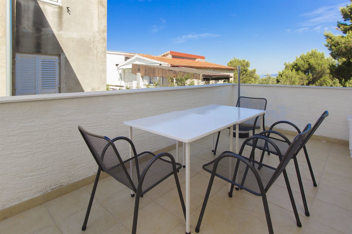 Um terraço com uma mesa branca e quatro cadeiras, uma parede branca e vista para edifícios e árvores.