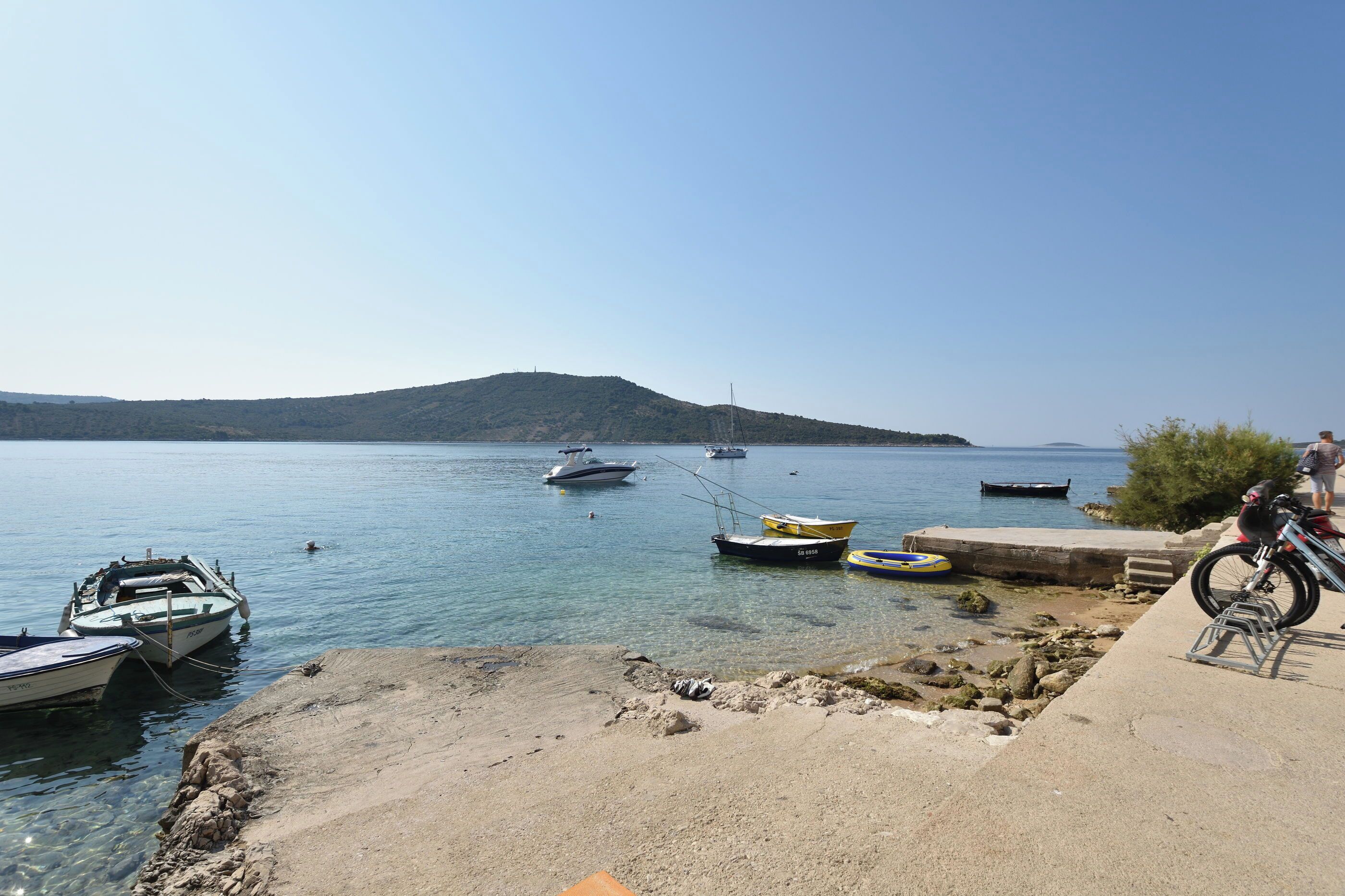 A coastal scene with boats docked near a sandy shore, a cyclist on the right, and a hilly landscape in the distance.