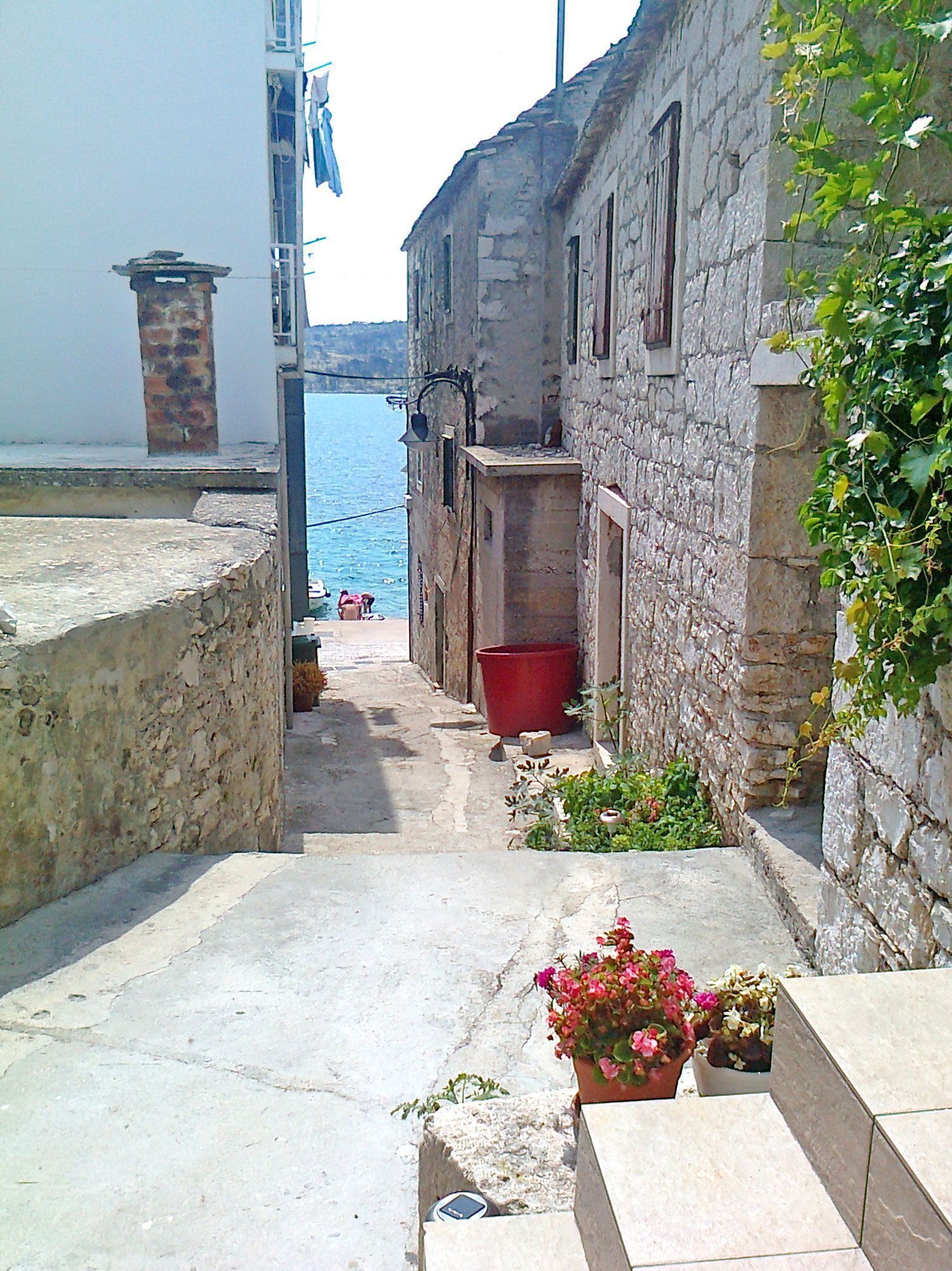 A narrow stone pathway leading to a body of water, flanked by old stone buildings with a red bucket and potted plants.