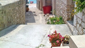 A narrow stone pathway leading to a body of water, flanked by old stone buildings with a red bucket and potted plants. - Apartment Milka (Primosten)