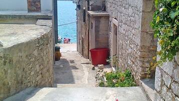 A narrow stone pathway leading to a body of water, flanked by old stone buildings with a red bucket and potted plants.