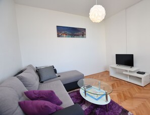 A modern living room with a sofa, a glass coffee table, a television, and a painting of a bridge. - Apartments Guzela (Primosten)