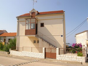 A two-story building with a red-tiled roof, a small balcony, and a gated entrance. - Apartments Guzela (Primosten)