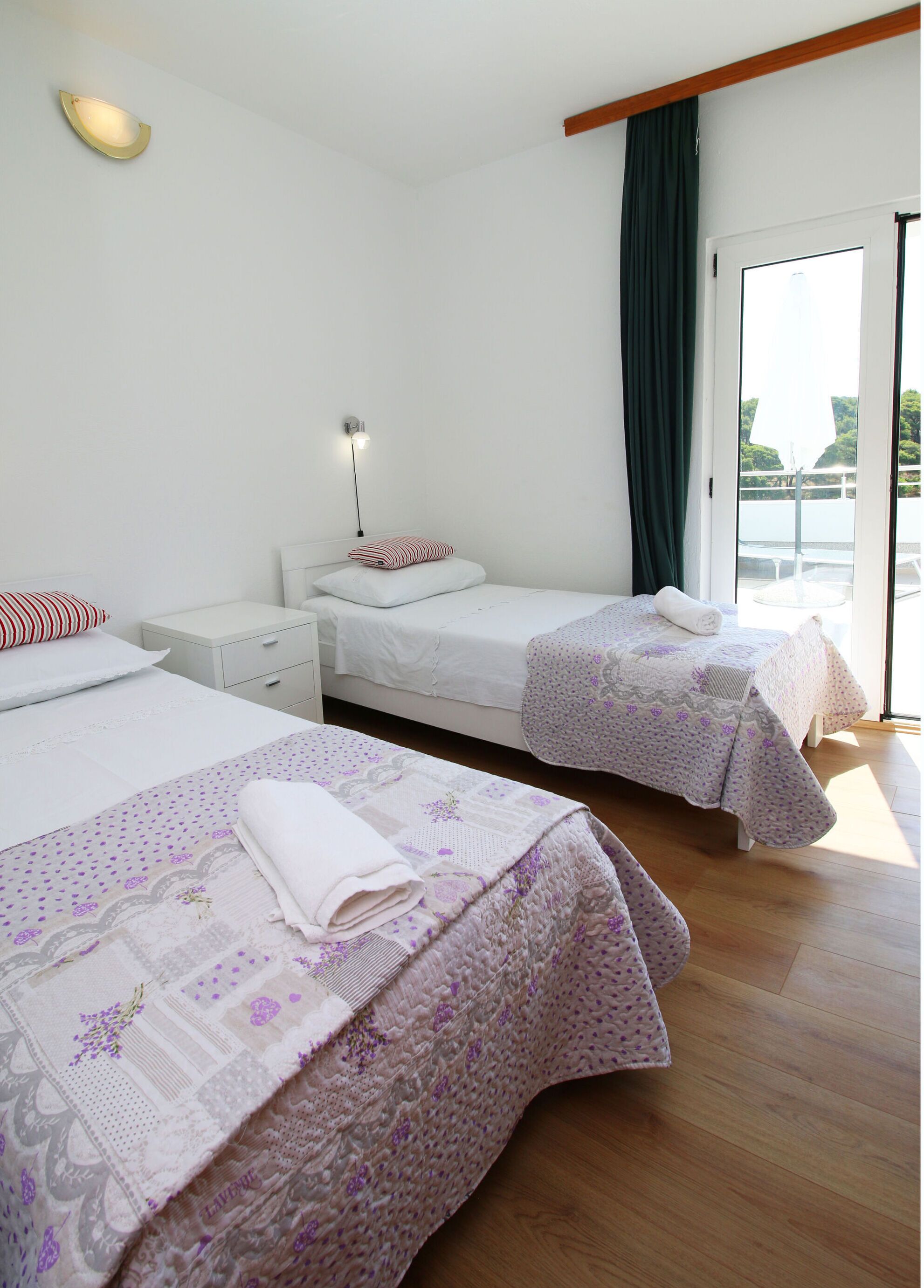 A hotel room with two beds, a nightstand, a lamp, and a view of the outdoors.