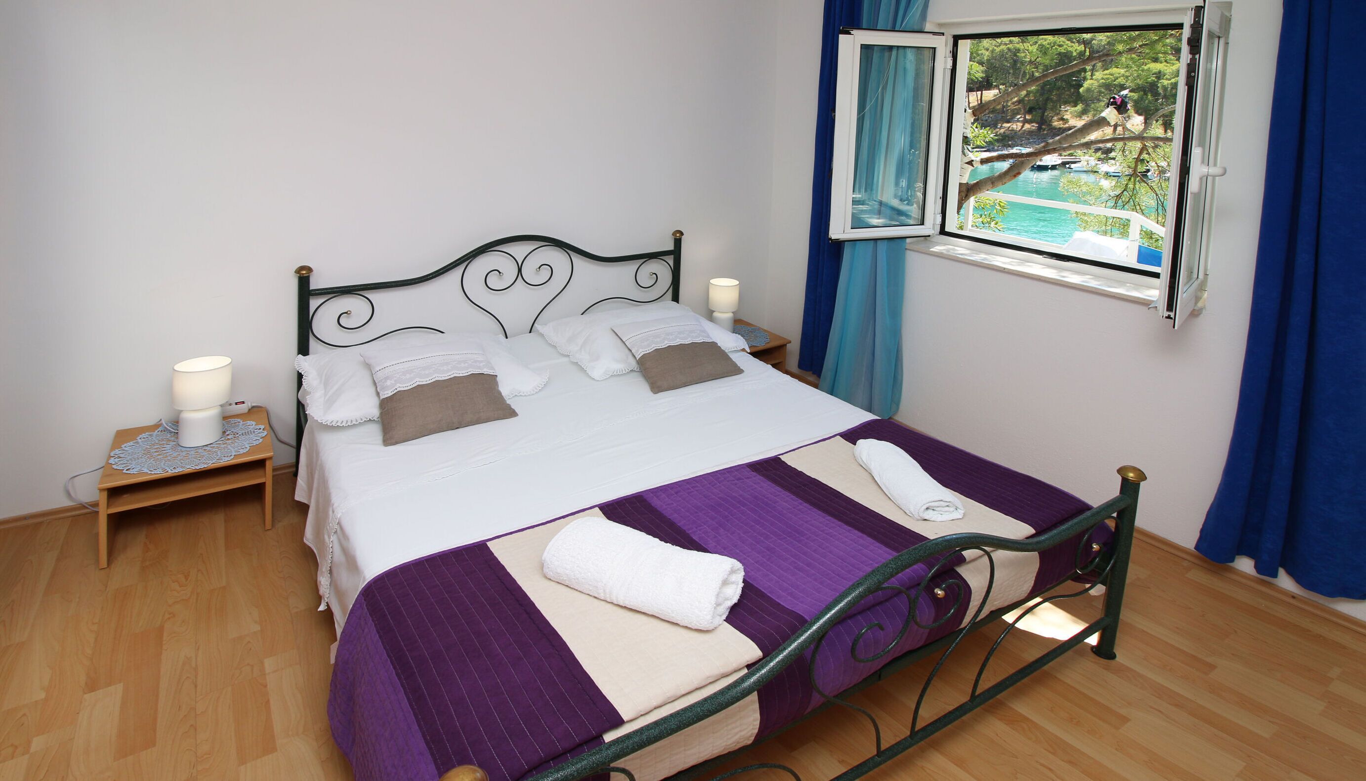 A neatly made bed with a purple and white striped bedspread, two pillows, and a folded white towel. There is a nightstand with a lamp and a window with a view of a body of water and trees.