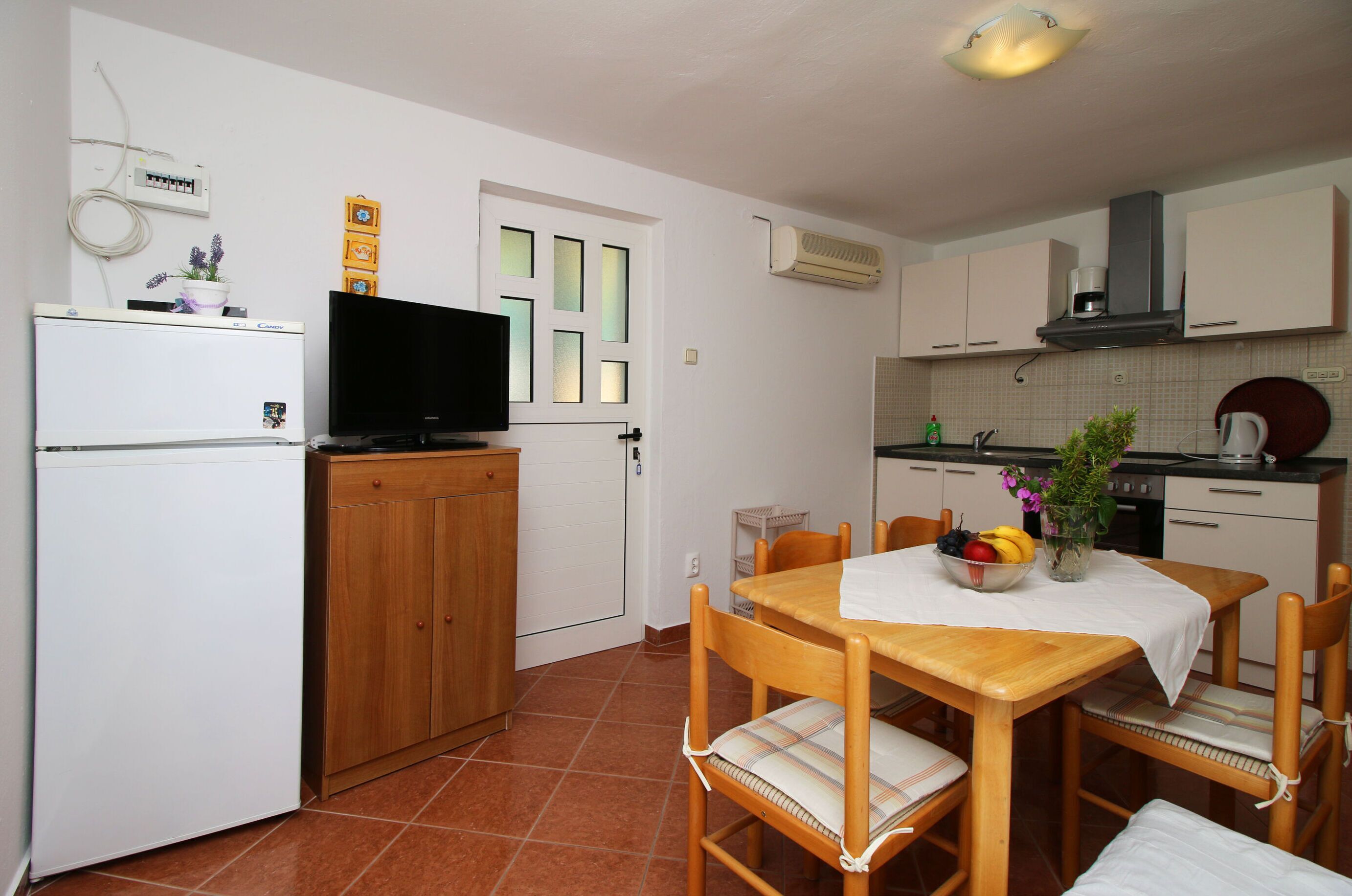 A compact kitchen with a dining area, white cabinets, a refrigerator, a television, and a small table with a fruit bowl and flowers.
