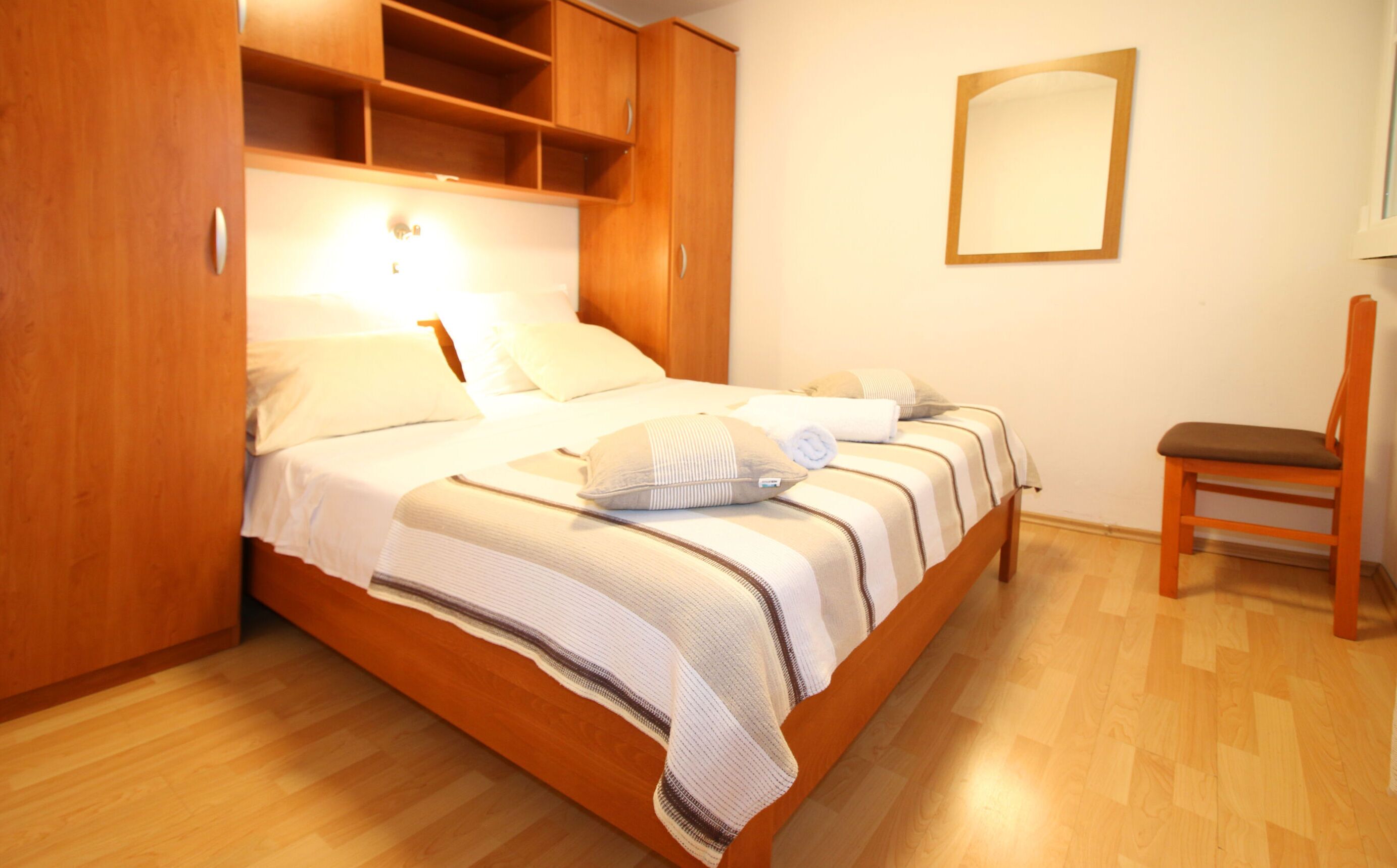 A neatly made bed with a striped blanket and pillows, a wooden wardrobe, a chair, and a mirror on the wall.