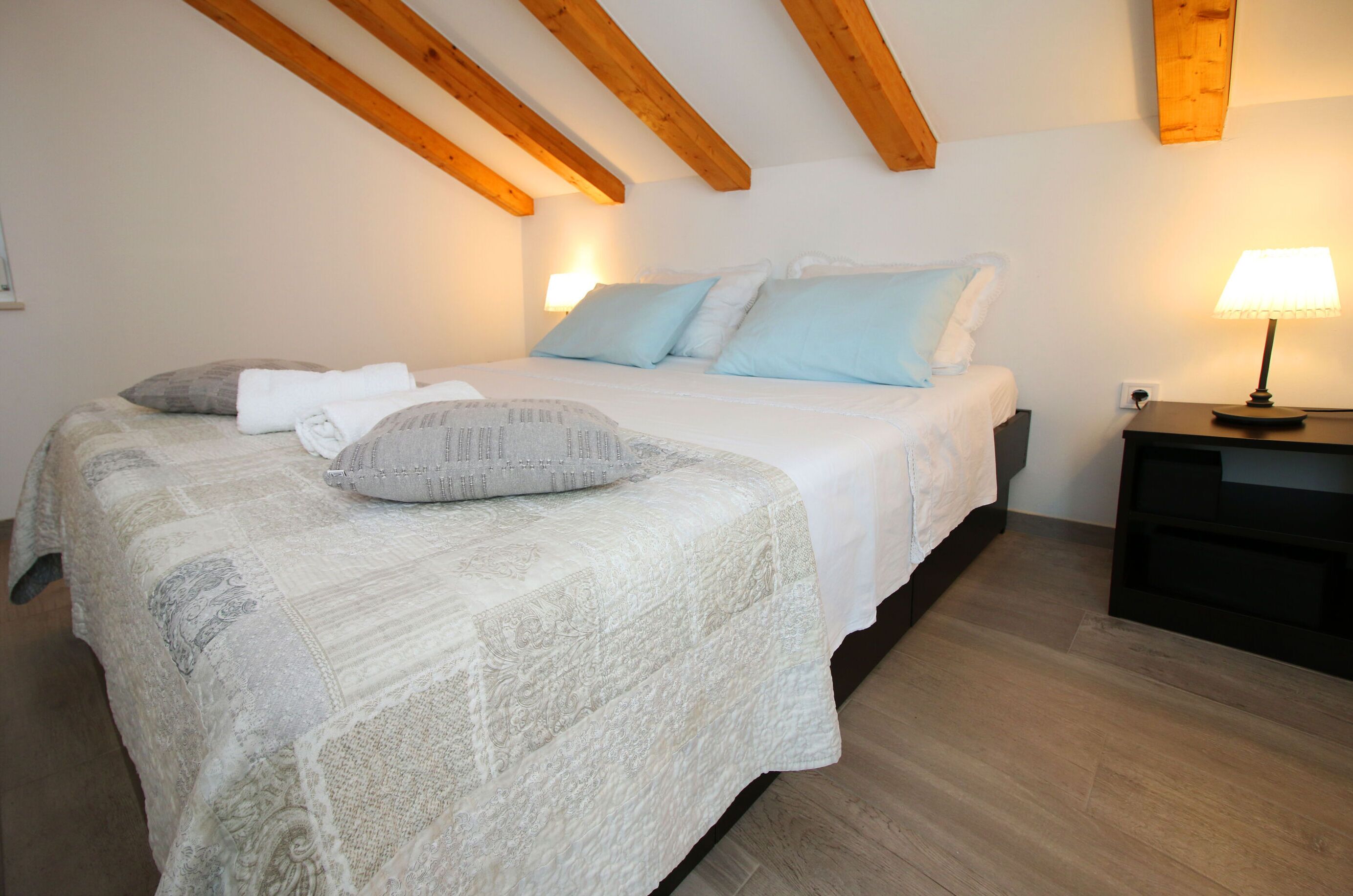 Un dormitorio con cama, almohadas, una mesita de noche con lámpara y vigas de madera en el techo.