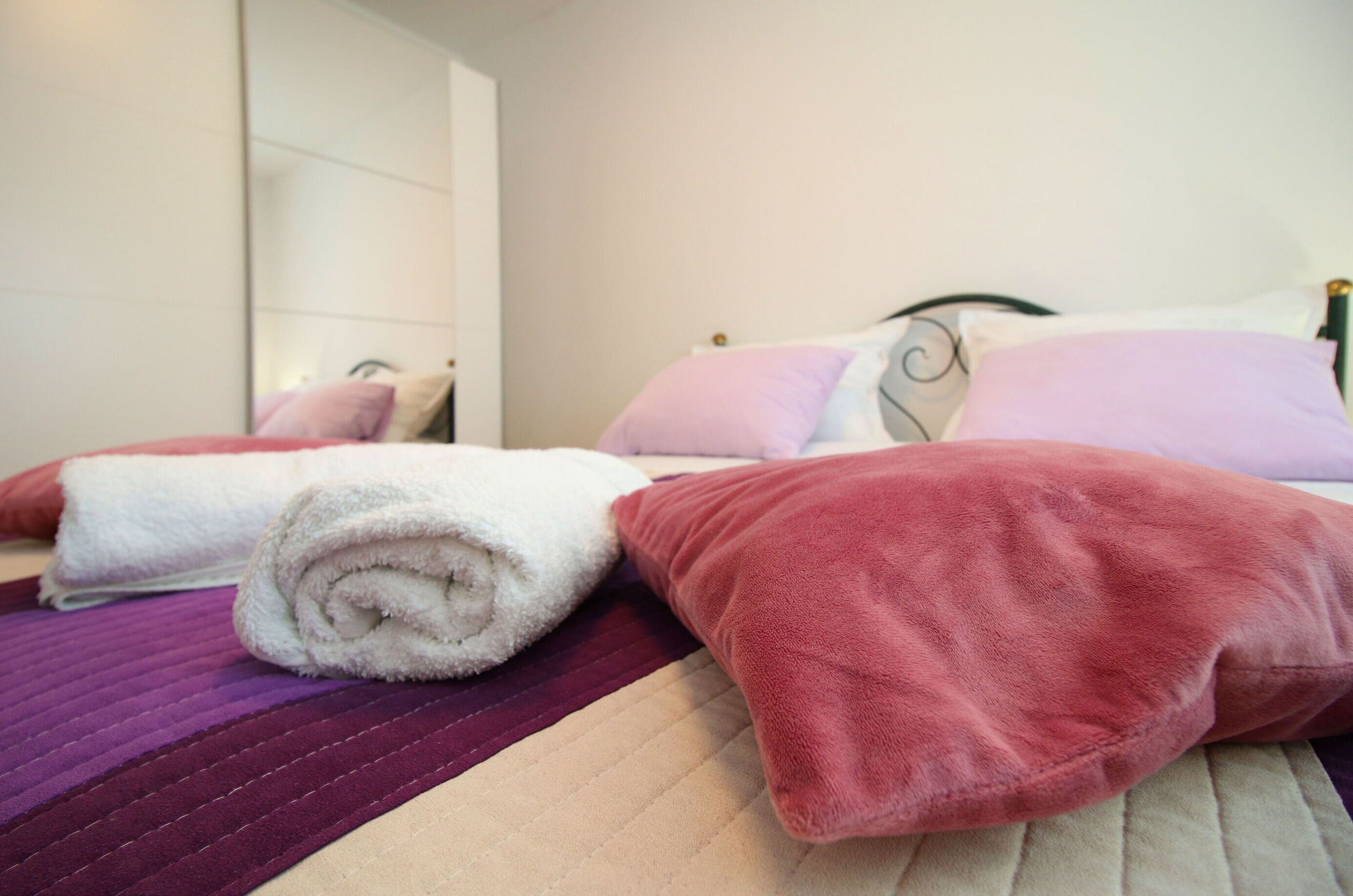 Una cama bien hecha con toallas blancas enrolladas, almohadas rosas y una colcha acolchada morada, junto a un cabecero con espejo.