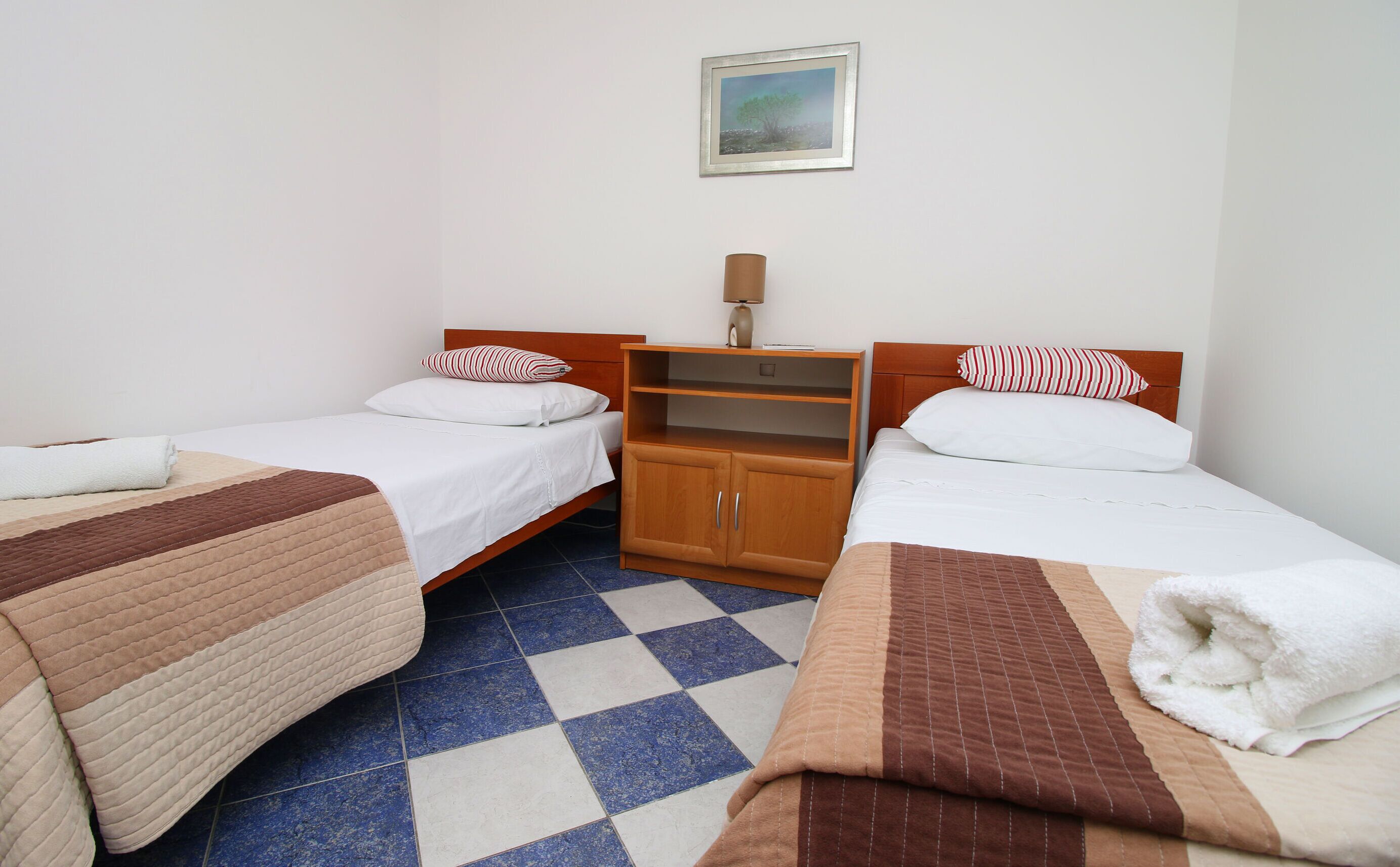 Una camera d'albergo con due letti singoli, un armadio in legno, un tavolino con una lampada e un quadro appeso al muro.