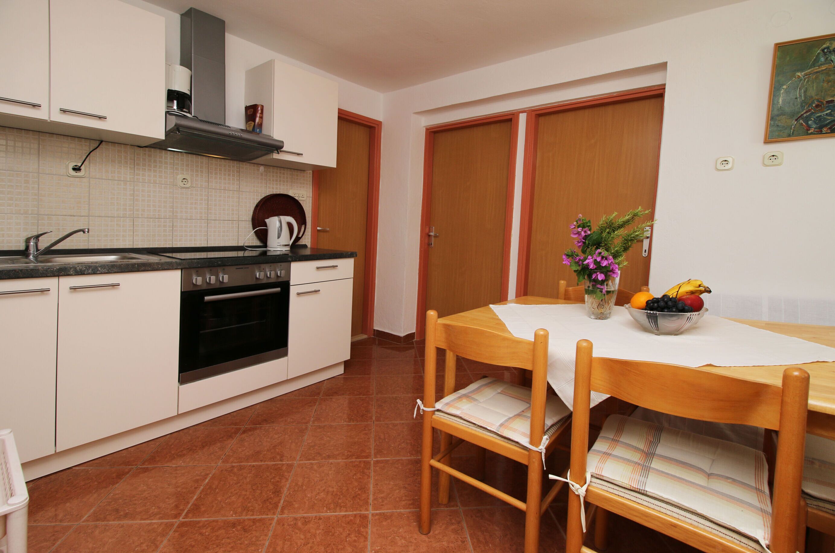A compact kitchen with white cabinets, a stove, and a sink. There's a dining table with chairs and a bowl of fruit.