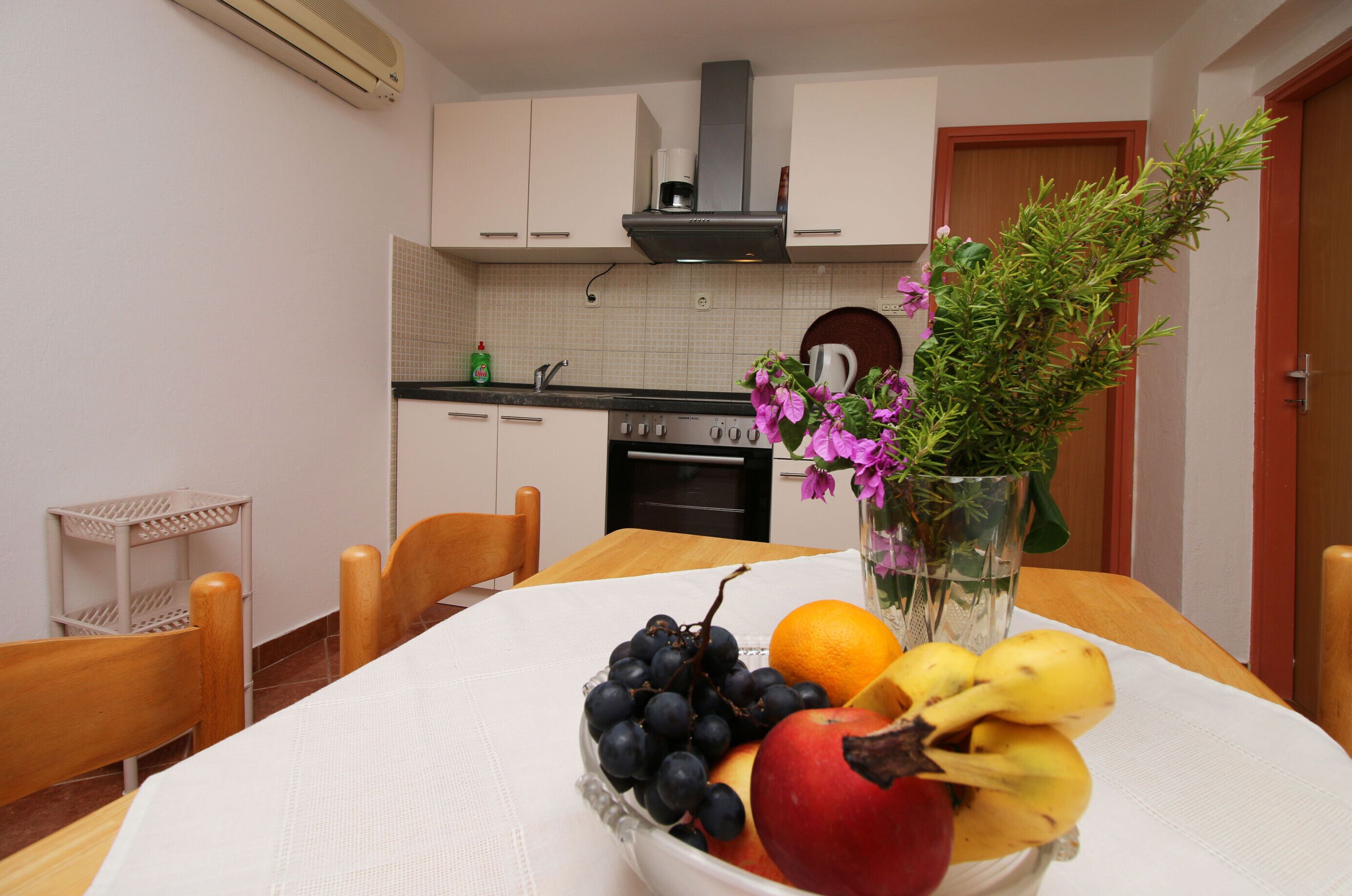 A dining table with a fruit bowl, a vase with flowers, and a kitchen in the background.