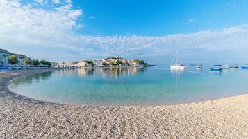Ein Kiesstrand mit klarem Wasser, einem Segelboot und Gebäuden im Hintergrund.
