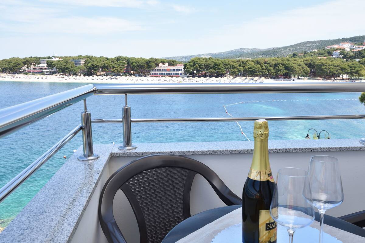 Un balcon avec une table, une bouteille de champagne et deux verres. La vue comprend une plage et une étendue d’eau.