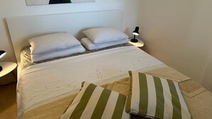 A neatly made bed with white linens and striped pillows, a bedside lamp, and a wall art piece above the bed. - Apartment Primosten Studio (Primosten)