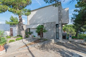 A modern building with stone walls, a flat roof, and a small garden area with potted plants and a palm tree. - Apartment Primosten Studio (Primosten)