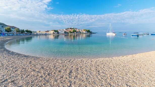 A pebbled beach with clear water, a sailboat, and buildings in the background. - Apartment Primosten Studio (Primosten)