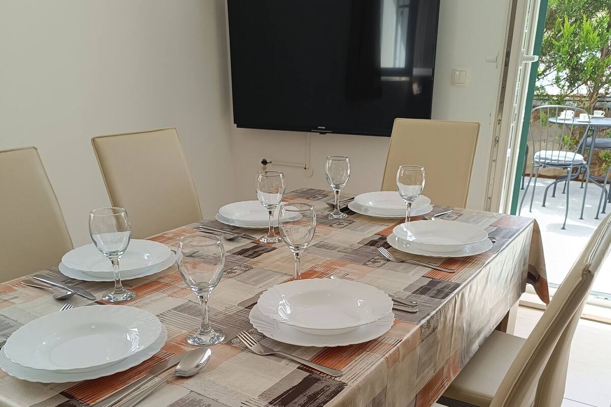 A dining table set for six with plates, glasses, and cutlery, a television above, and a view of the outdoors through a glass door.
