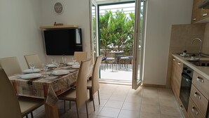A dining area with a table set for four, a television above the table, and a kitchen with a sink and cabinets. - Apartments Antonia (Primosten)