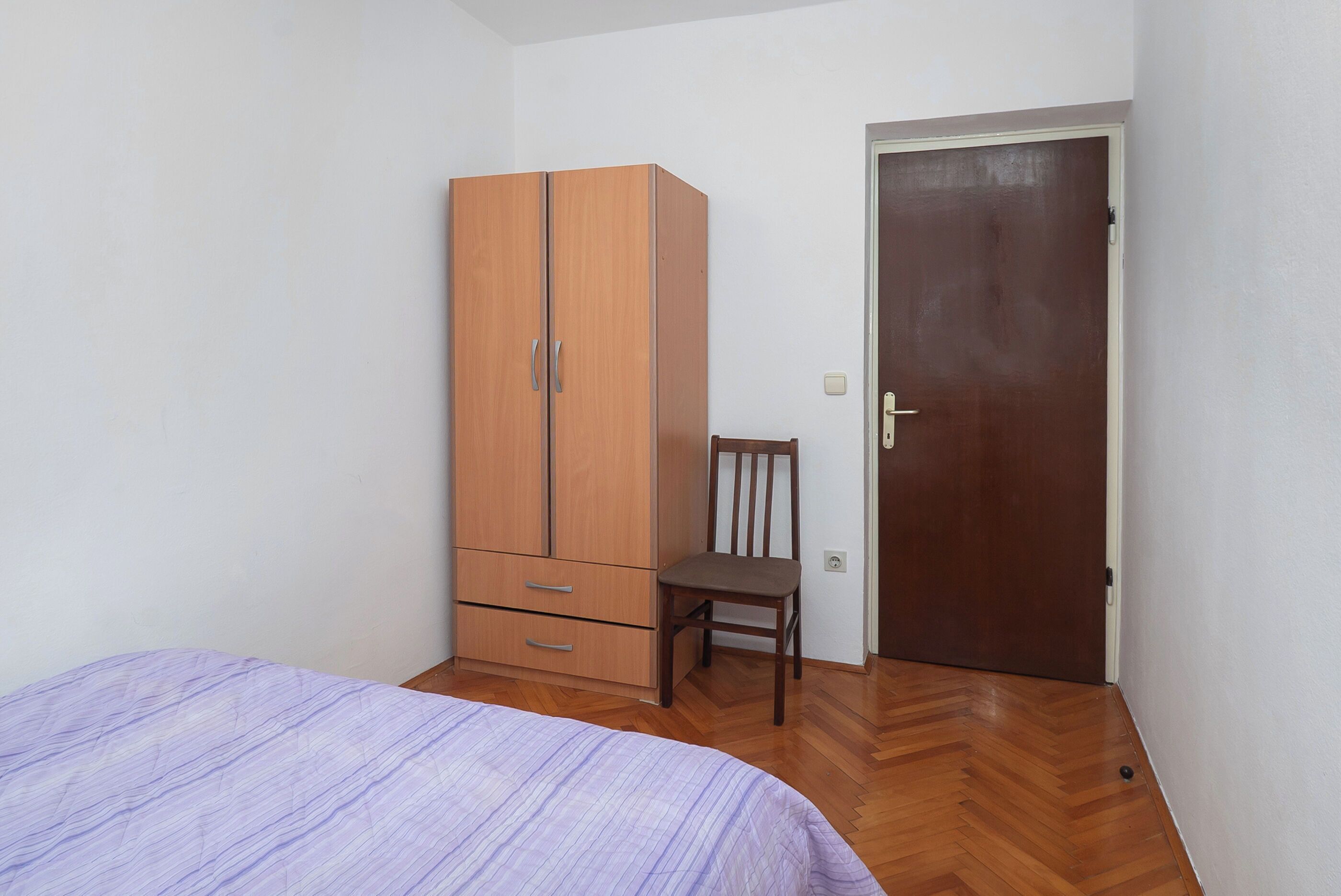 A bedroom with a bed, wooden wardrobe, chair, and a door.