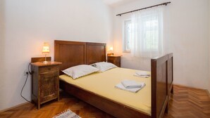 Un dormitorio pequeño y limpio con una cama de madera, dos mesitas de noche con lámparas, un ventanal con cortinas traslúcidas y suelo de madera.