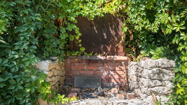 Un muro de piedra con chimenea de ladrillo, rodeado de follaje verde.