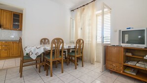 Un comedor con una mesa lista para cuatro personas, sillas de madera, un televisor sobre un mueble de madera, y una zona de cocina con armarios de madera.