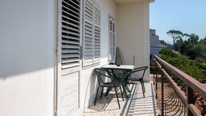 Un balcón con una mesa y sillas, contraventanas blancas, y vistas a árboles y edificios.