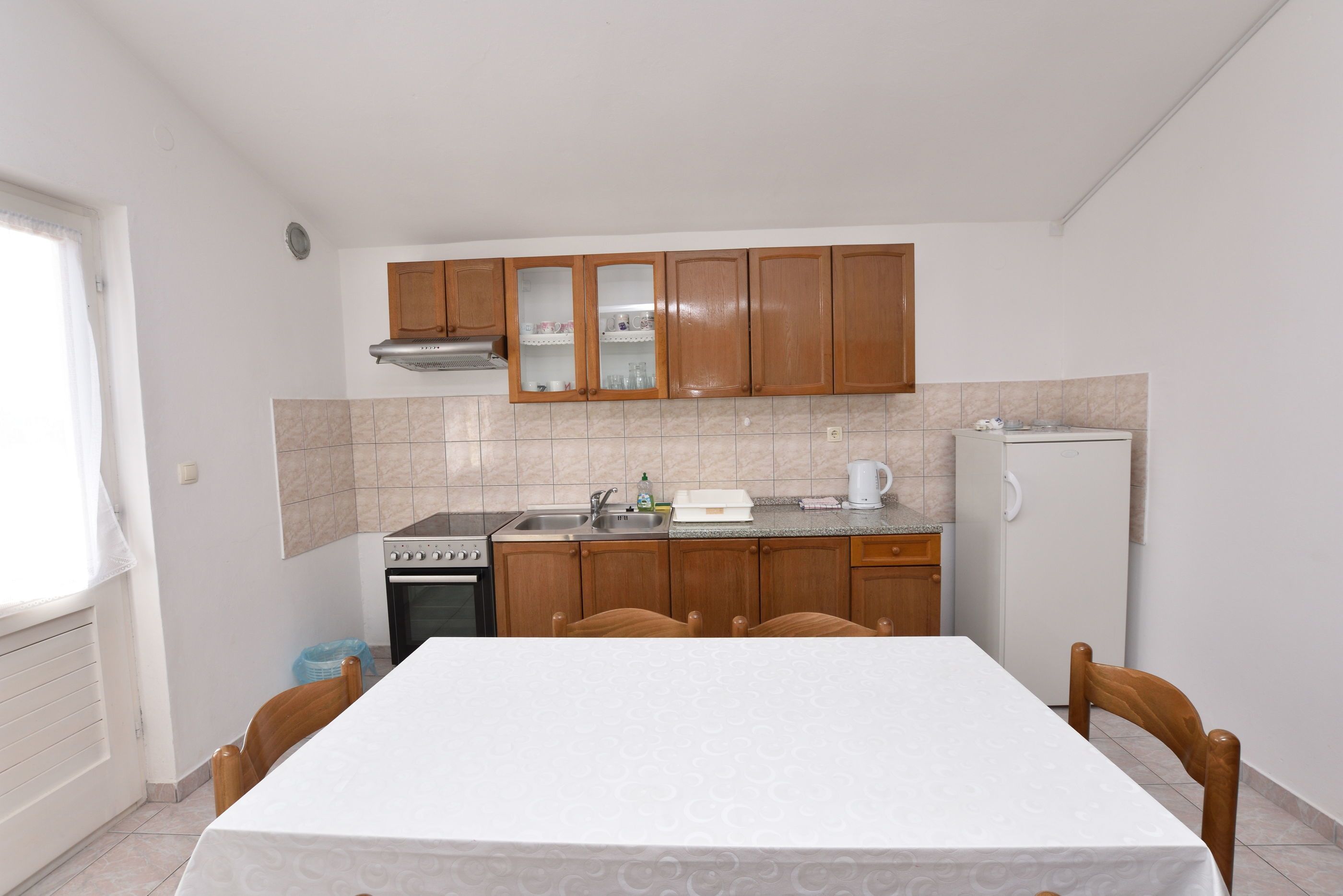 A compact kitchen with wooden cabinets, a stove, sink, and a refrigerator. There is a dining table with chairs in the center.