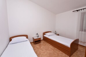 A small, clean room with two single beds, wooden headboards, a nightstand with a lamp, and a window with a sheer curtain. - Apartment Damir (Primosten)