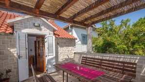 A covered outdoor seating area with a wooden bench and a table, a stone building with a tiled roof, and a potted plant. - House Sanda (Kastel Novi)
