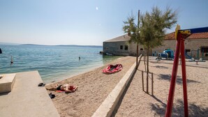 A beach with a sandy area, a building with a tiled roof, and a playground with red equipment. - House Sanda (Kastel Novi)