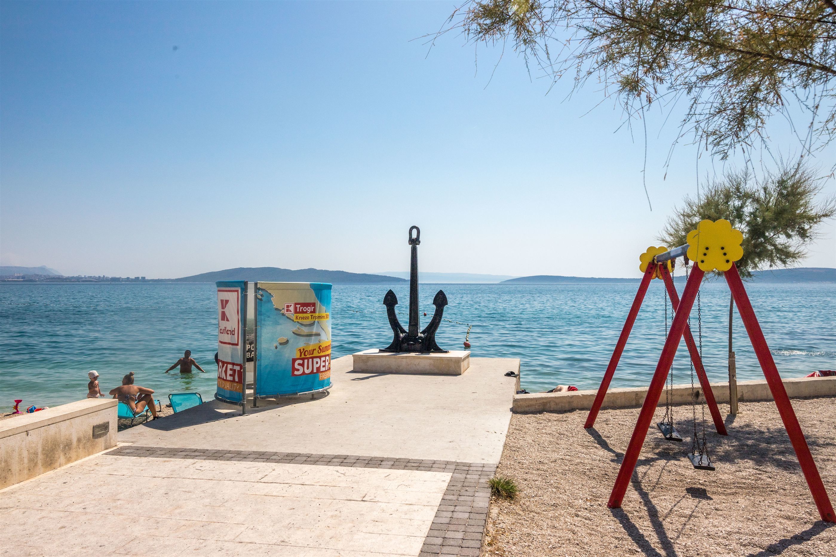 A seaside promenade with a swing set, a statue, and a kiosk.