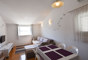 Ett litet, väl upplyst rum med en soffa, ett matbord, en tv och ett fönster med gardiner.