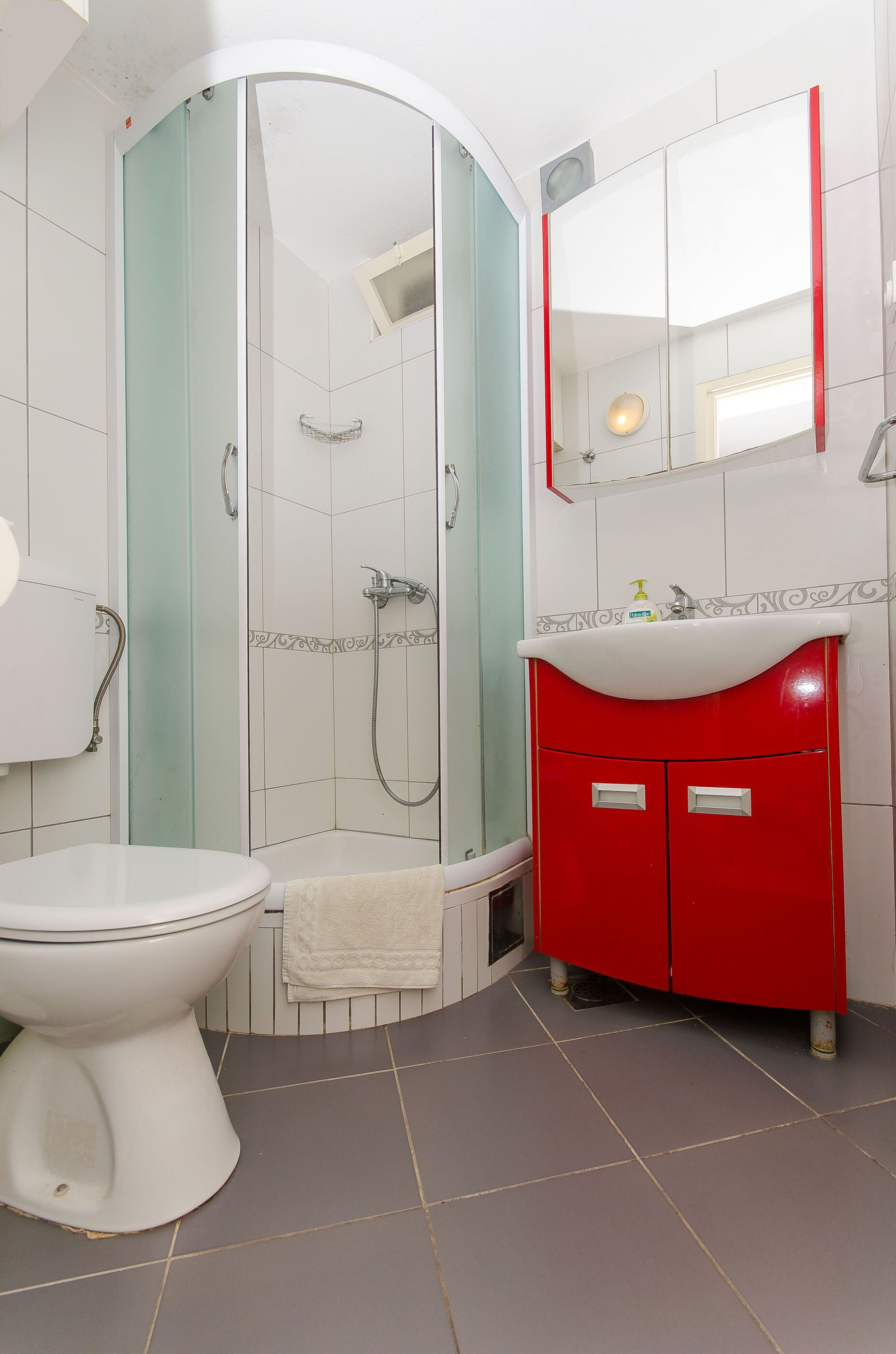 A bathroom with a white toilet, a glass-enclosed shower, a red vanity with a white sink, and a mirror above the vanity.