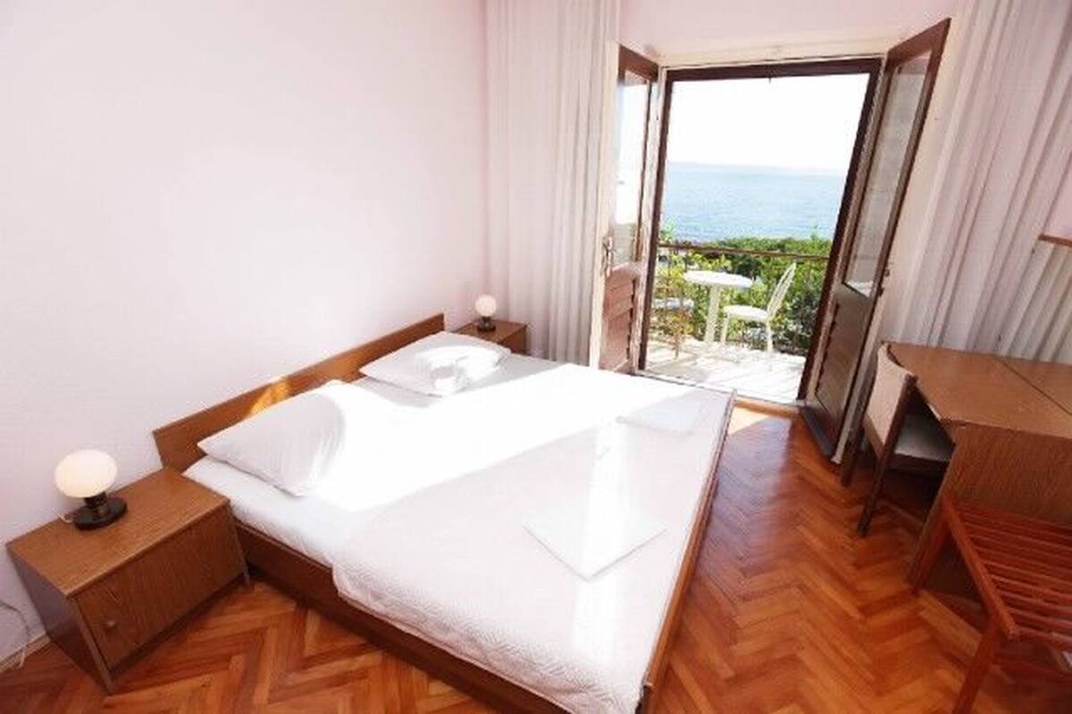 A hotel room with a bed, a nightstand, a desk, and a view of the sea through an open door.