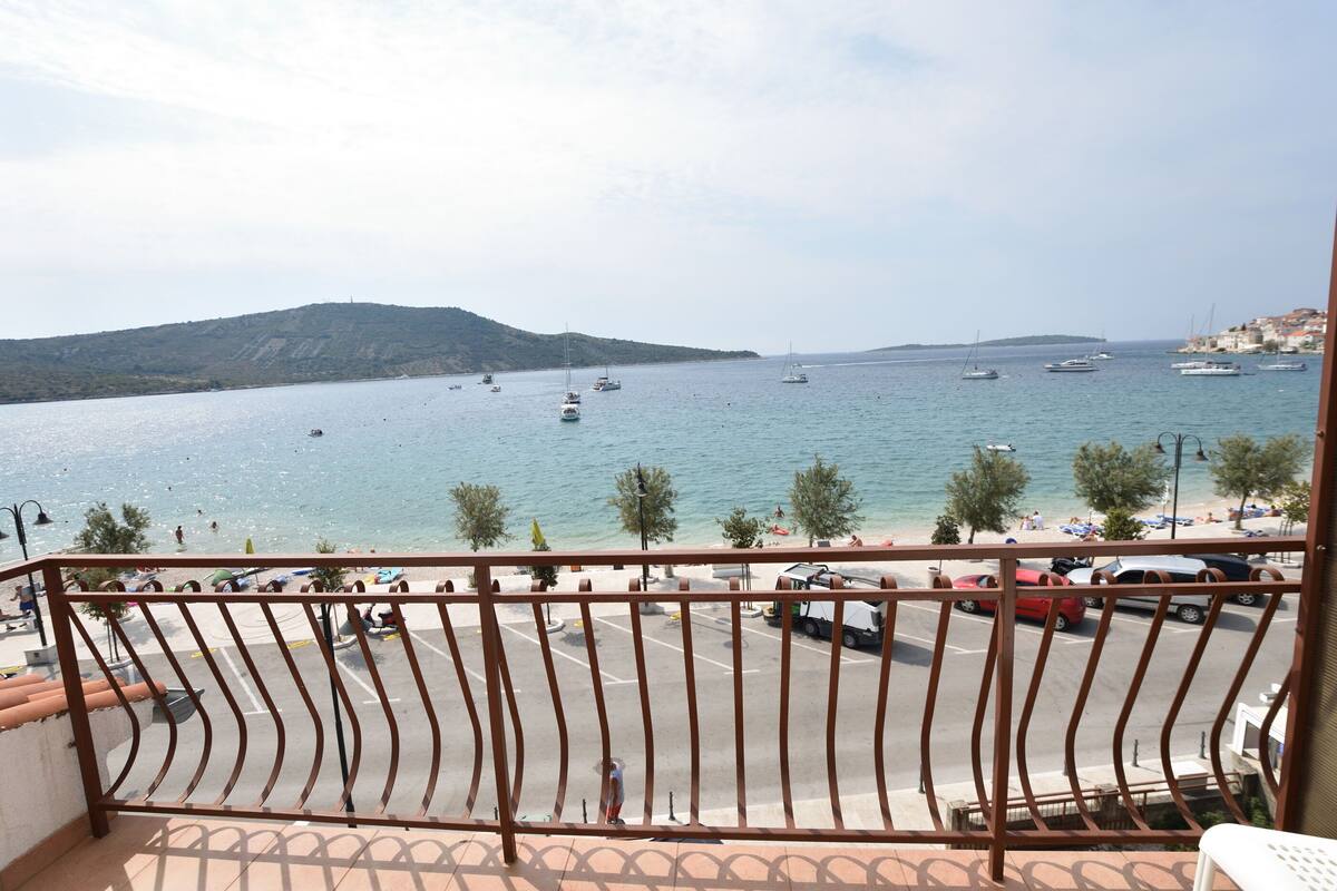 Un balcon avec vue sur une mer calme, des bateaux et un parking où sont garées des voitures et un camion.