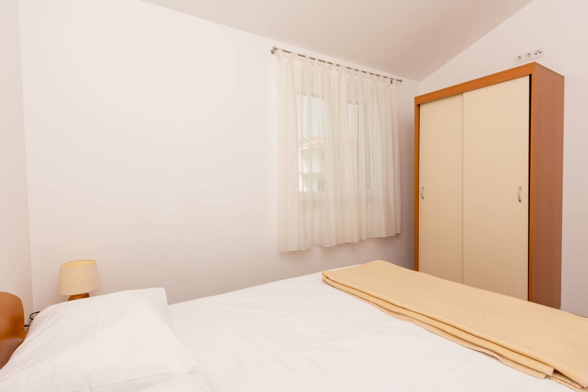 Une chambre à coucher avec un lit, une armoire en bois, une fenêtre avec des rideaux légers et une lampe de chevet.