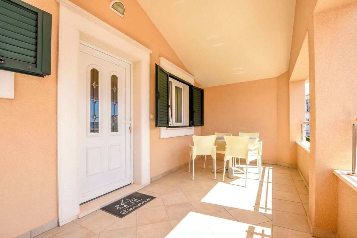 Un couloir avec une porte blanche, des volets verts et un sol carrelé. La pièce contient deux chaises et une table.
