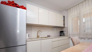 A modern kitchen with white cabinets, a stainless steel refrigerator, a wooden dining table, and a window with sheer curtains. - Apartments Frane (Primosten)